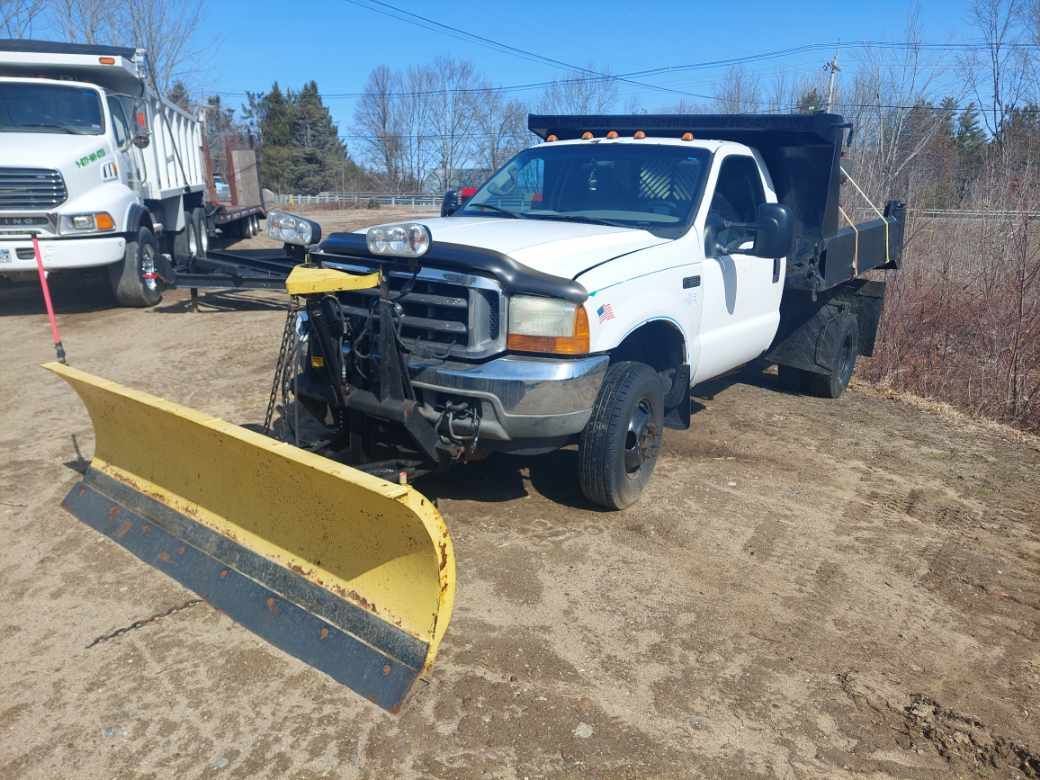 A white truck with a yellow snow plow on the front is parked on a dirt road.