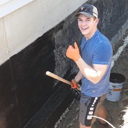 A Happy Worker Applying Waterproof Paint on a House Foundation