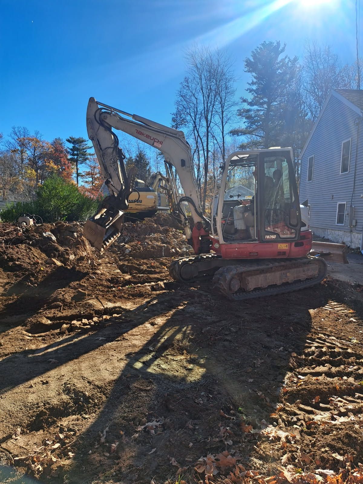 Excavator digging a trench next to a house under a sunny, blue sky.