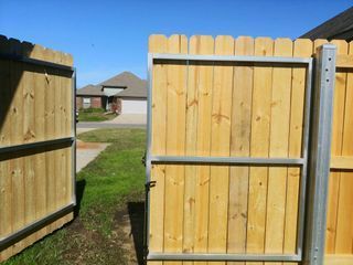 Wooden fence gate open, revealing a green yard and a house under a blue sky.