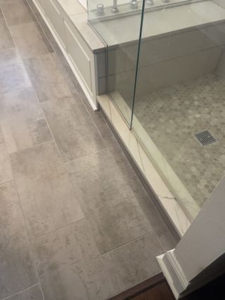 Tiled bathroom floor leading to a glass shower with pebble floor, marble trim, and white baseboards.