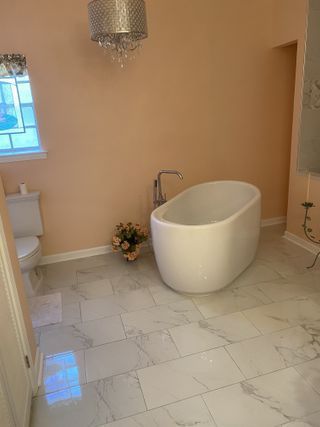 White soaking tub in a peach-colored bathroom with marble tile floors and a crystal chandelier.