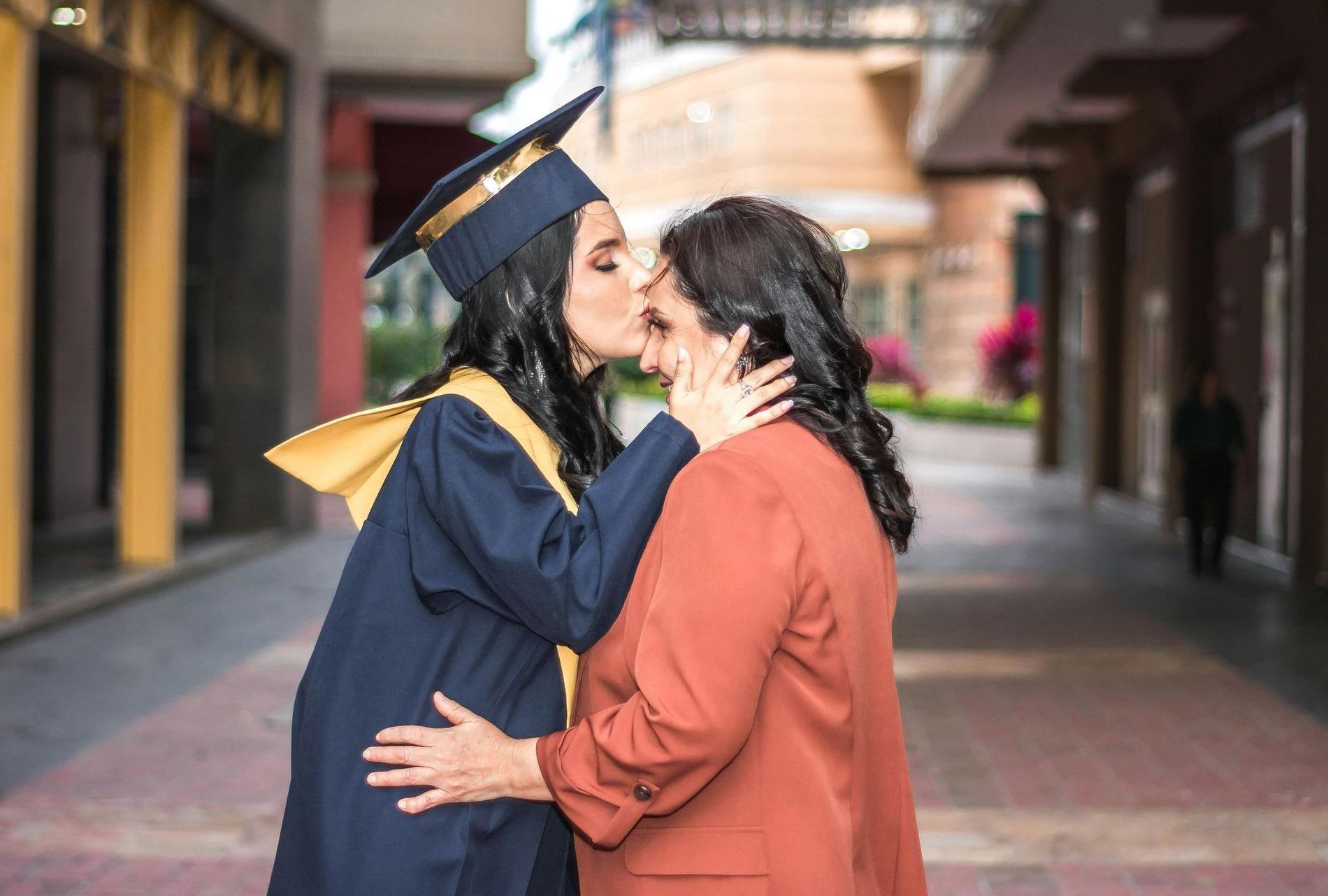 A woman in a graduation cap and gown is kissing her mother on the cheek.