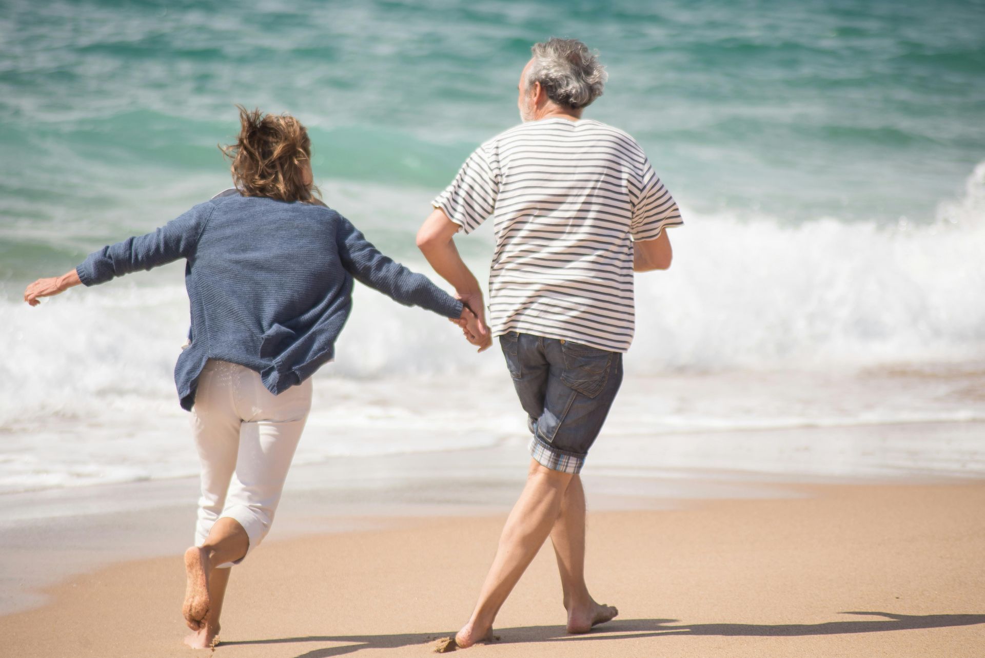 An elderly couple is walking on the beach holding hands.