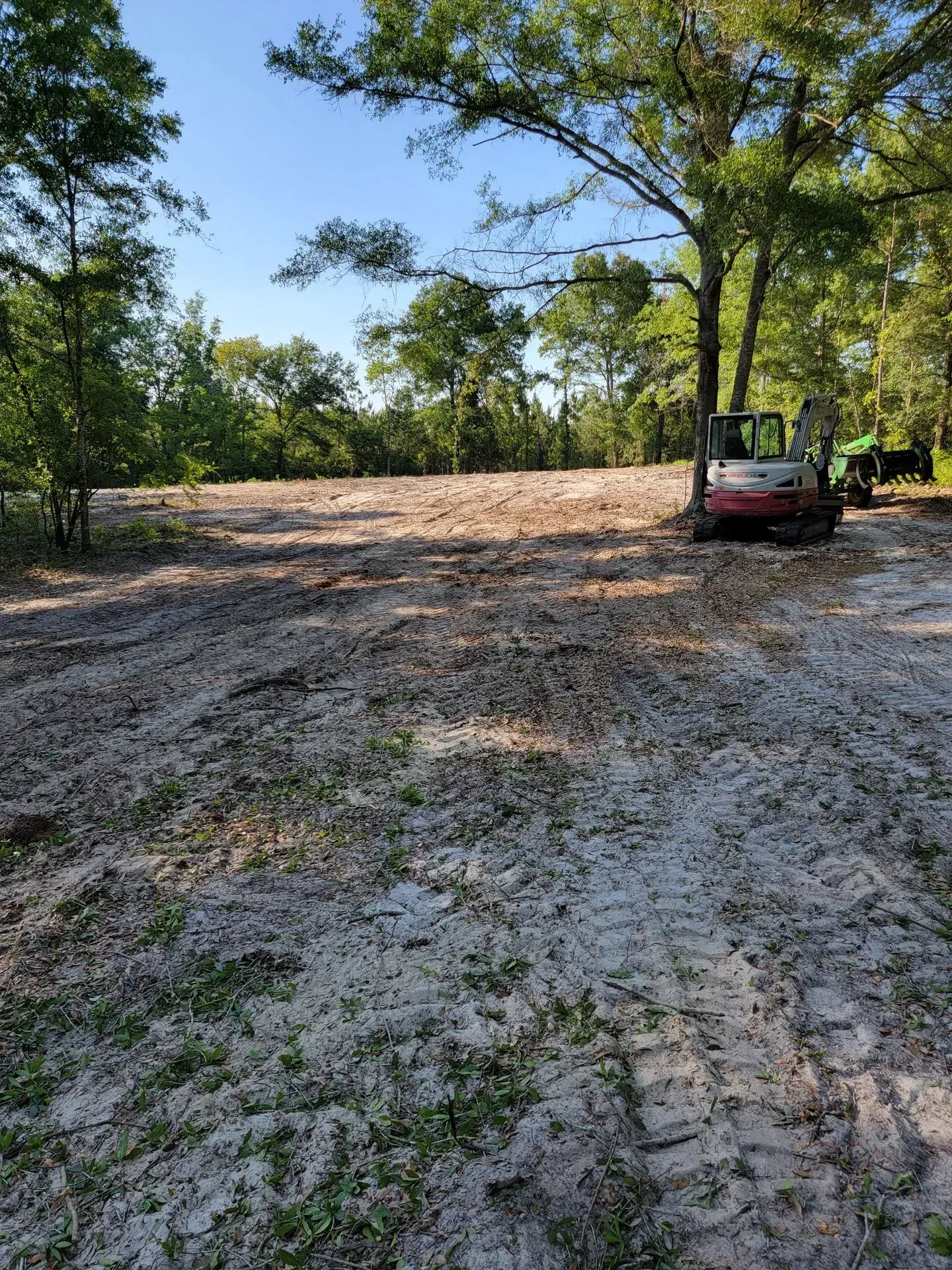 Cleared land with an excavator and tractor visible, surrounded by trees under a blue sky.
