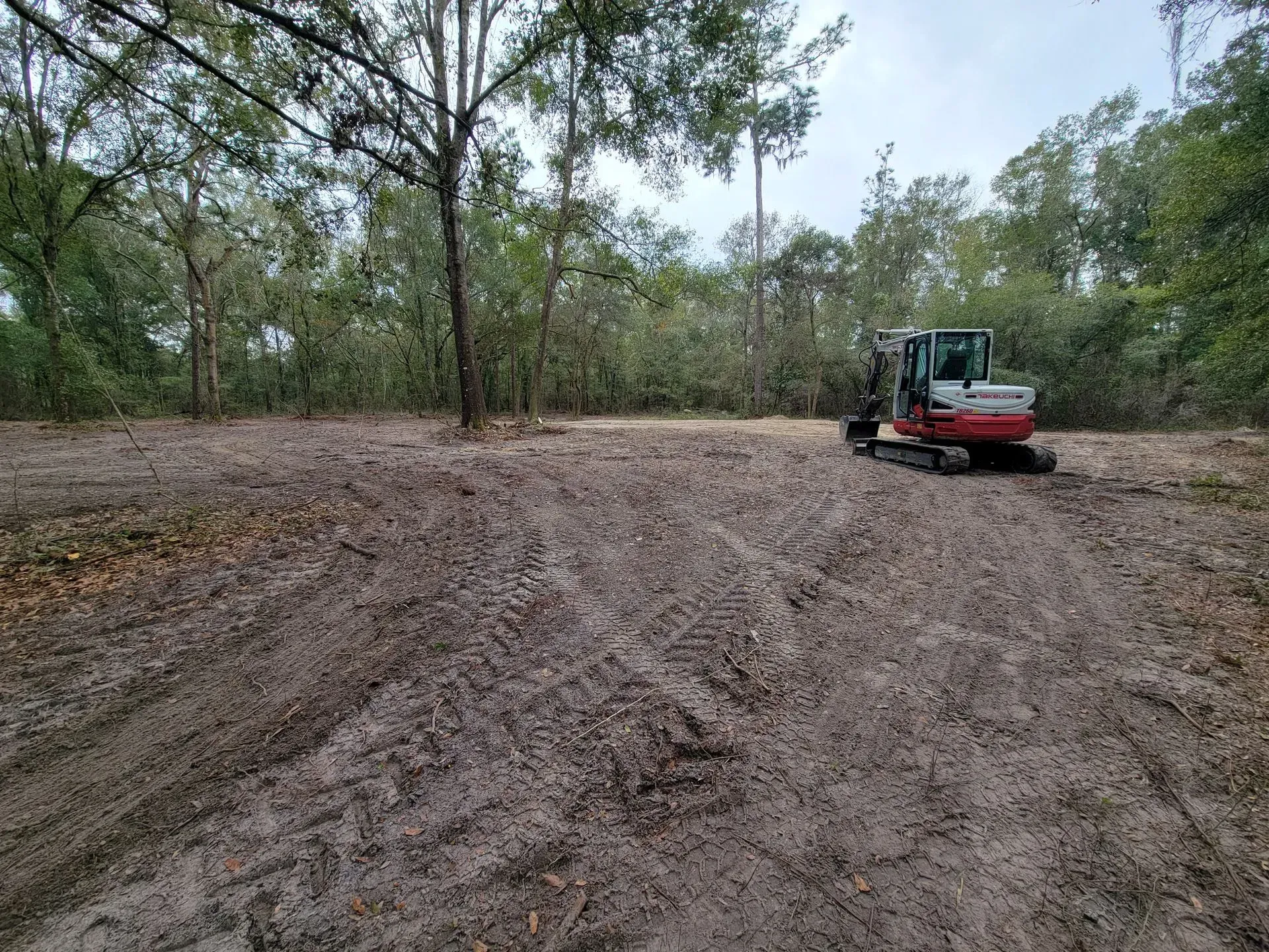 A small excavator on a cleared dirt lot in a wooded area. The sky is overcast.