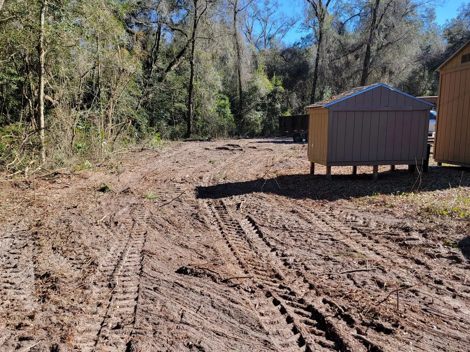 Muddy clearing with tire tracks, sheds, and trees in a wooded area.