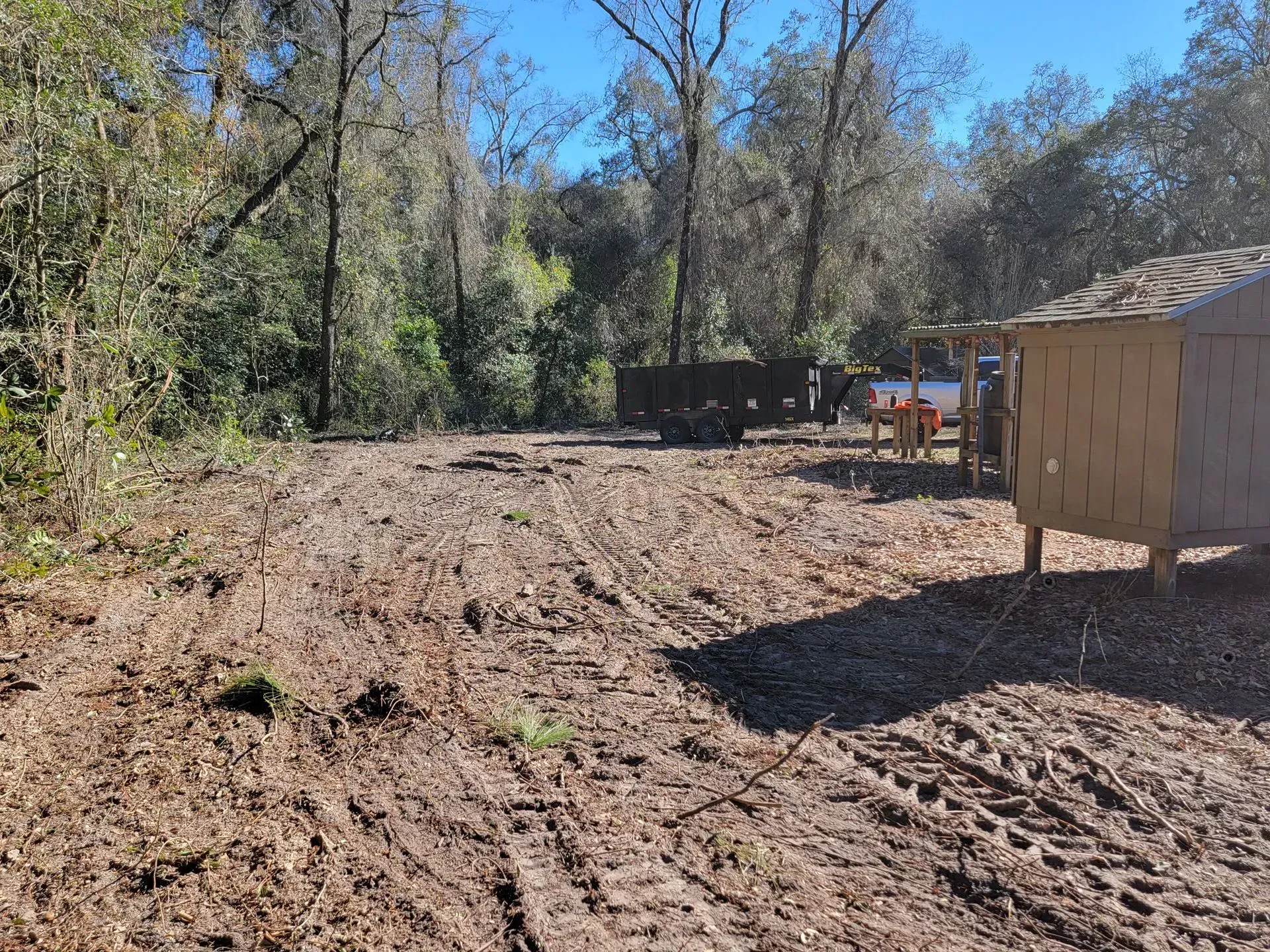 Cleared dirt area with tire tracks, small wooden structure, and trees in background.