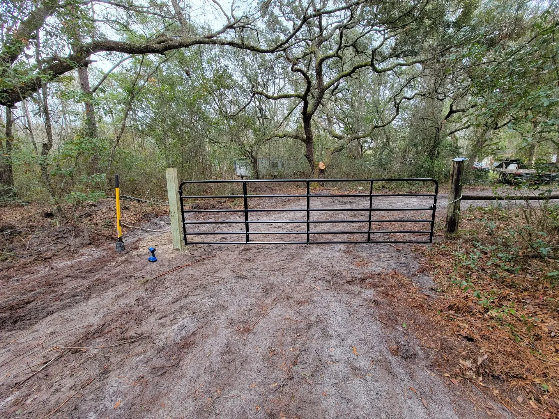 Black metal gate blocks a dirt road entrance, surrounded by trees.