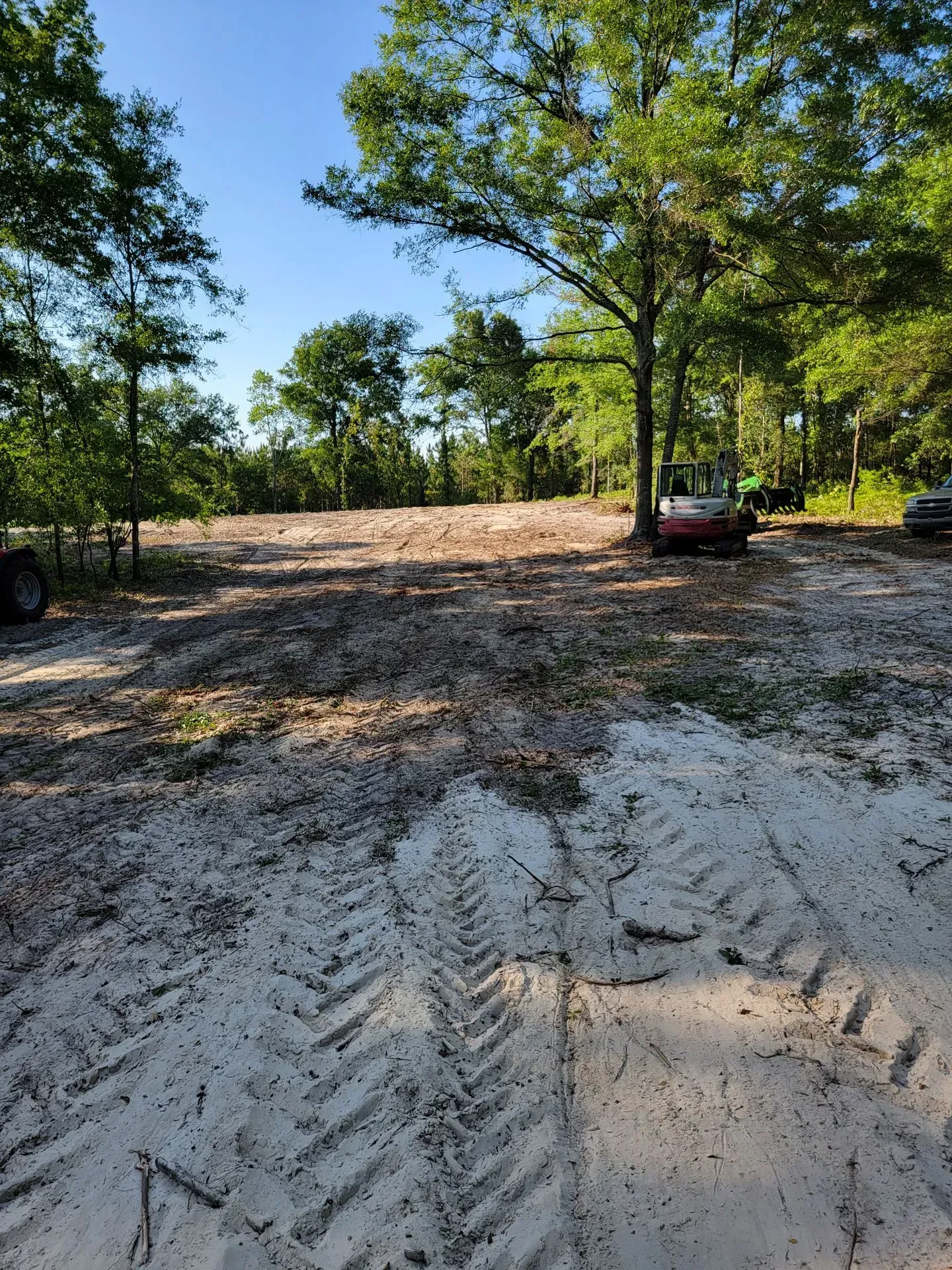 Sandy clearing with tractor tracks; trees surround a cleared area, vehicles parked nearby.