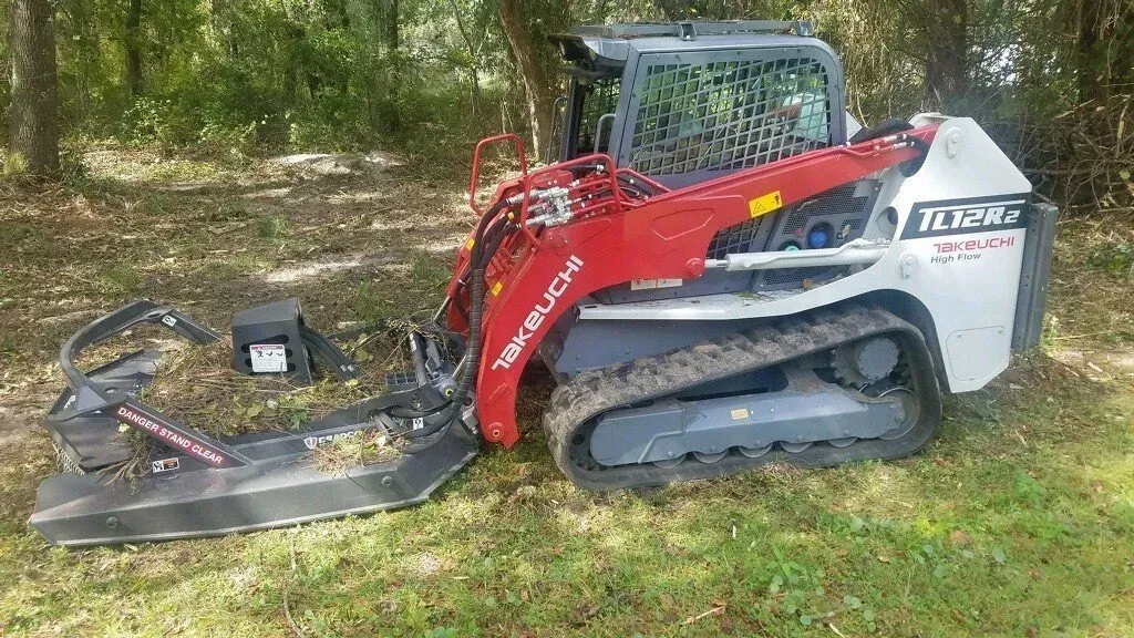Red and gray compact track loader with brush cutter attachment on grass.