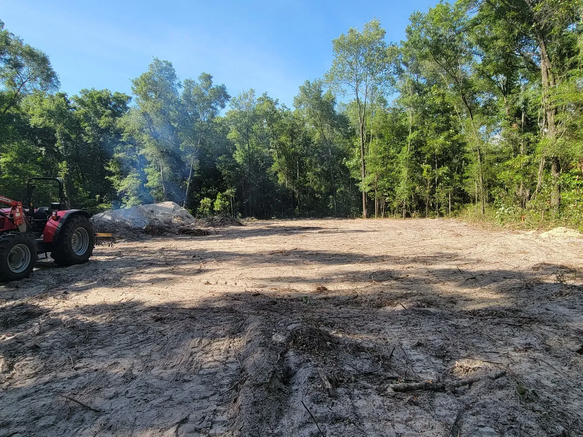 Tractor near cleared land with smoking pile of debris, surrounded by trees under a blue sky.