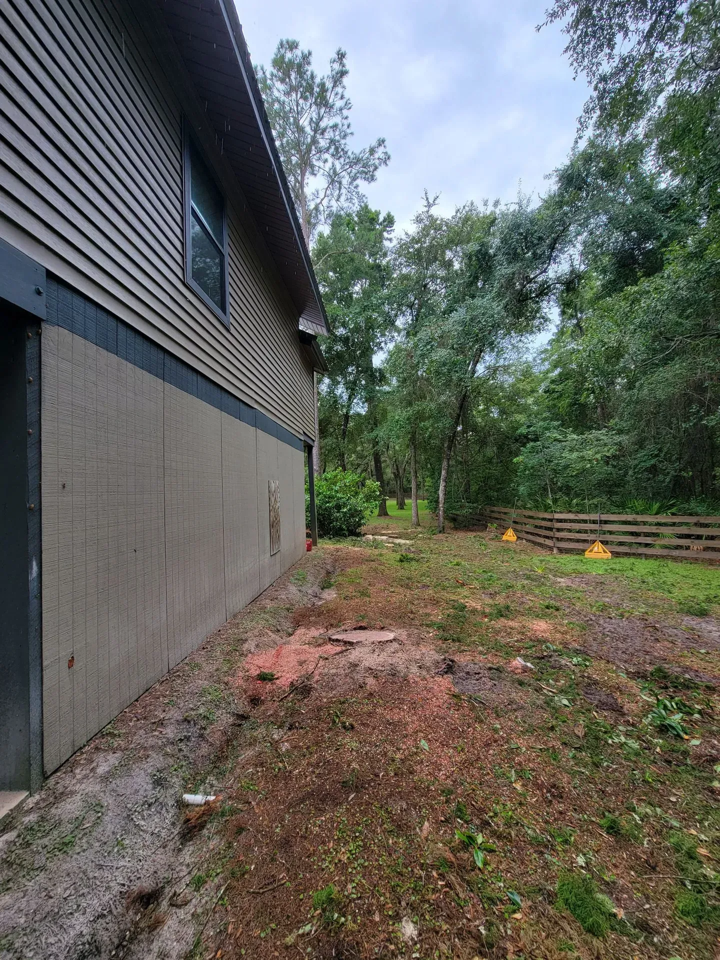 Side view of a house with a dirt yard, leading to a wooden fence and trees. Gray and brown colors dominate.