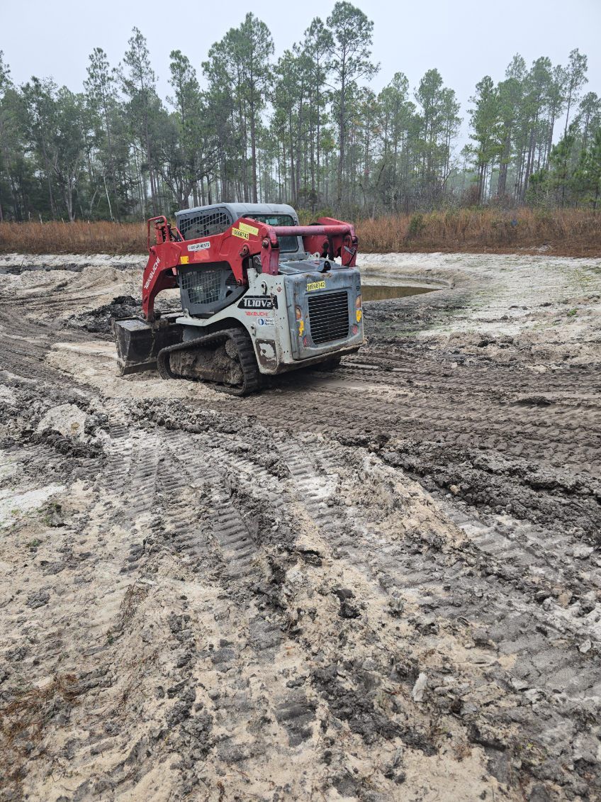 Small, tracked excavator working in muddy, cleared land, with trees in the background.