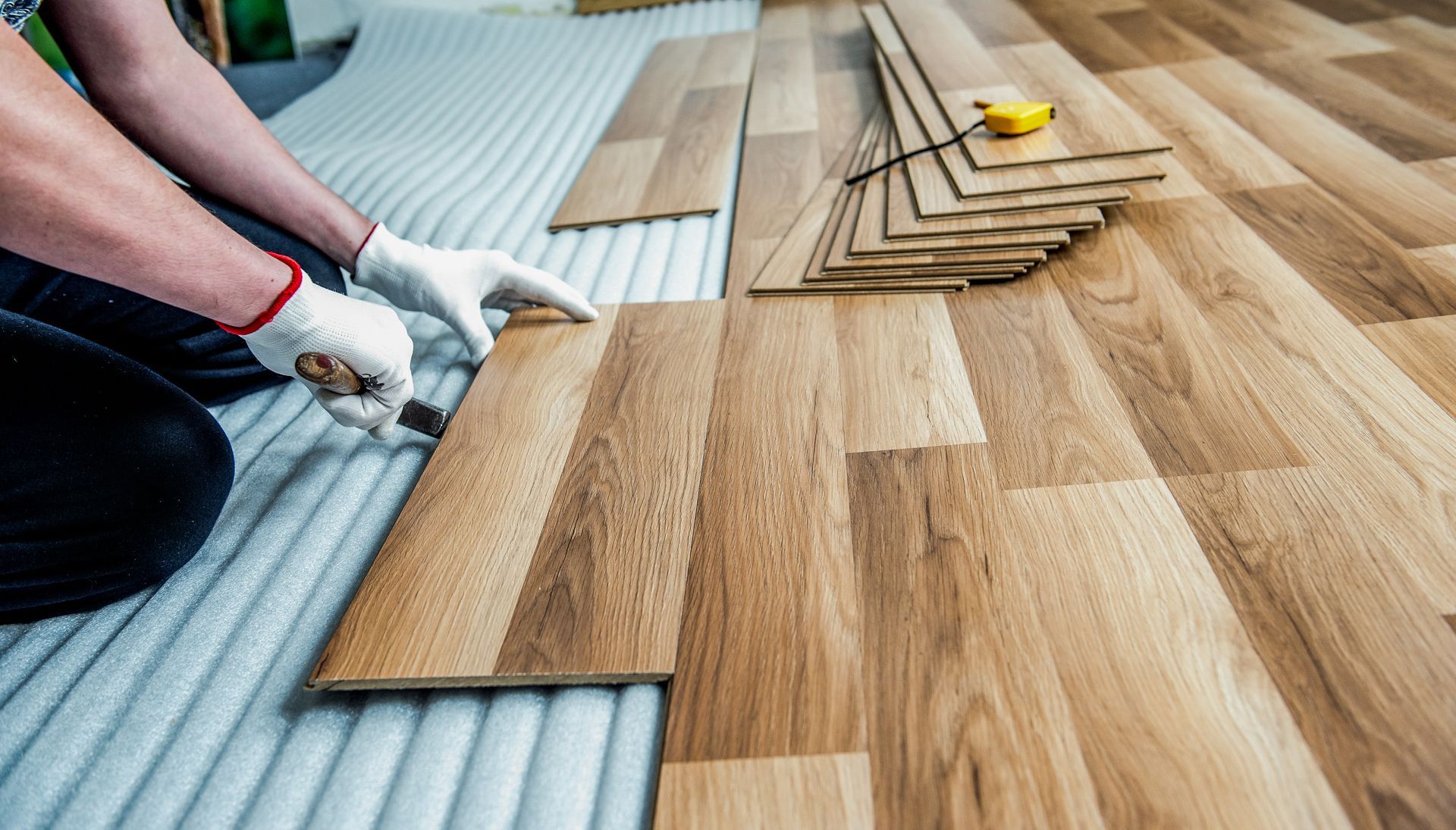 Person installing wood flooring with a utility knife, wearing gloves, indoors.