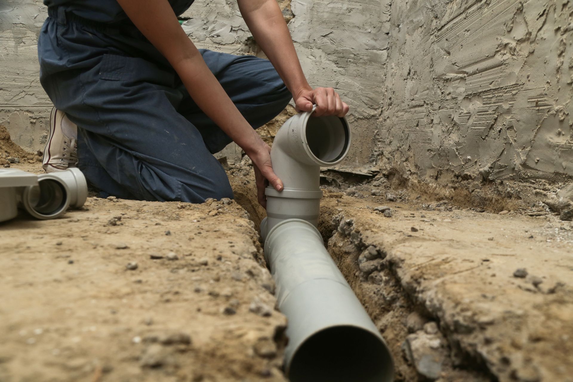 Person kneeling, connecting grey PVC pipe and elbow in a trench. Concrete wall background.