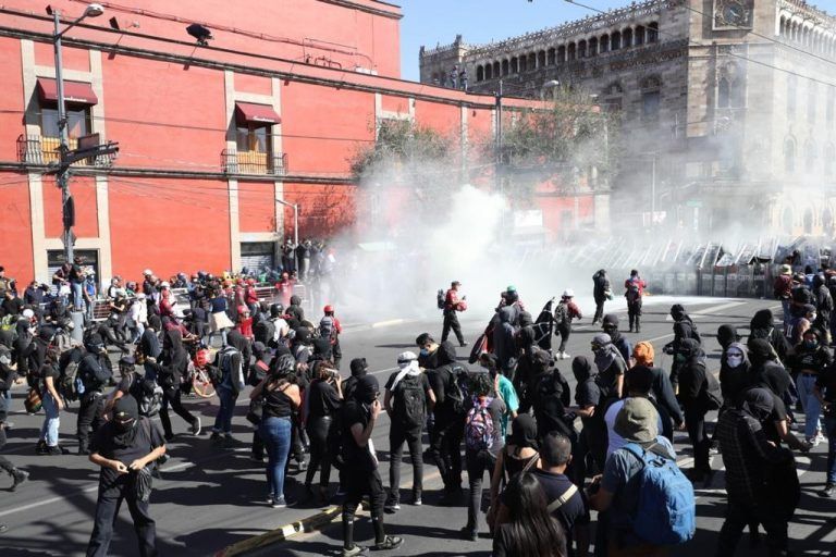 Protesters in black face water cannons in a city square, red building in background.