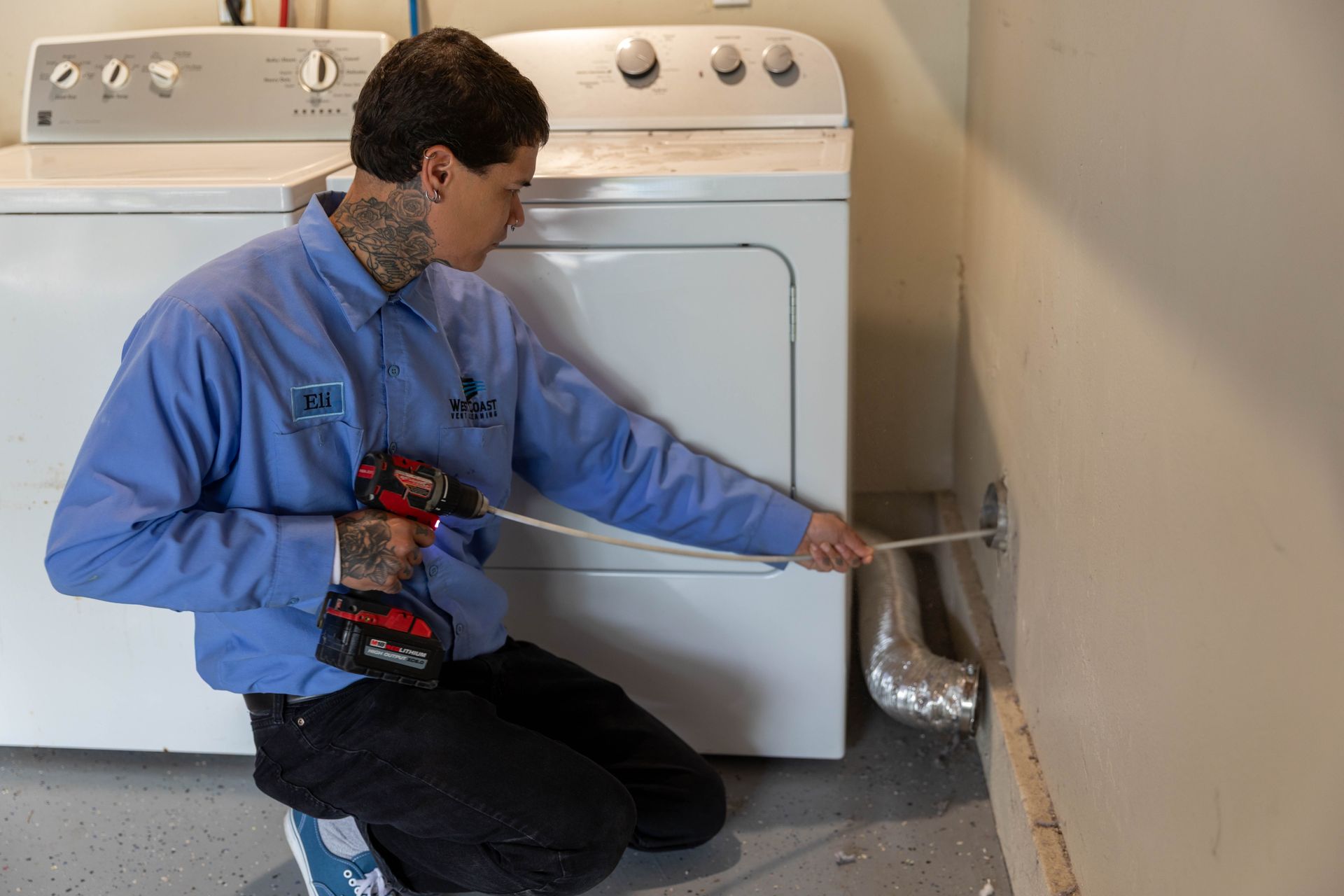 A technician in a blue uniform uses a drill attachment to clean a dryer vent inside a wall behind laundry appliances.