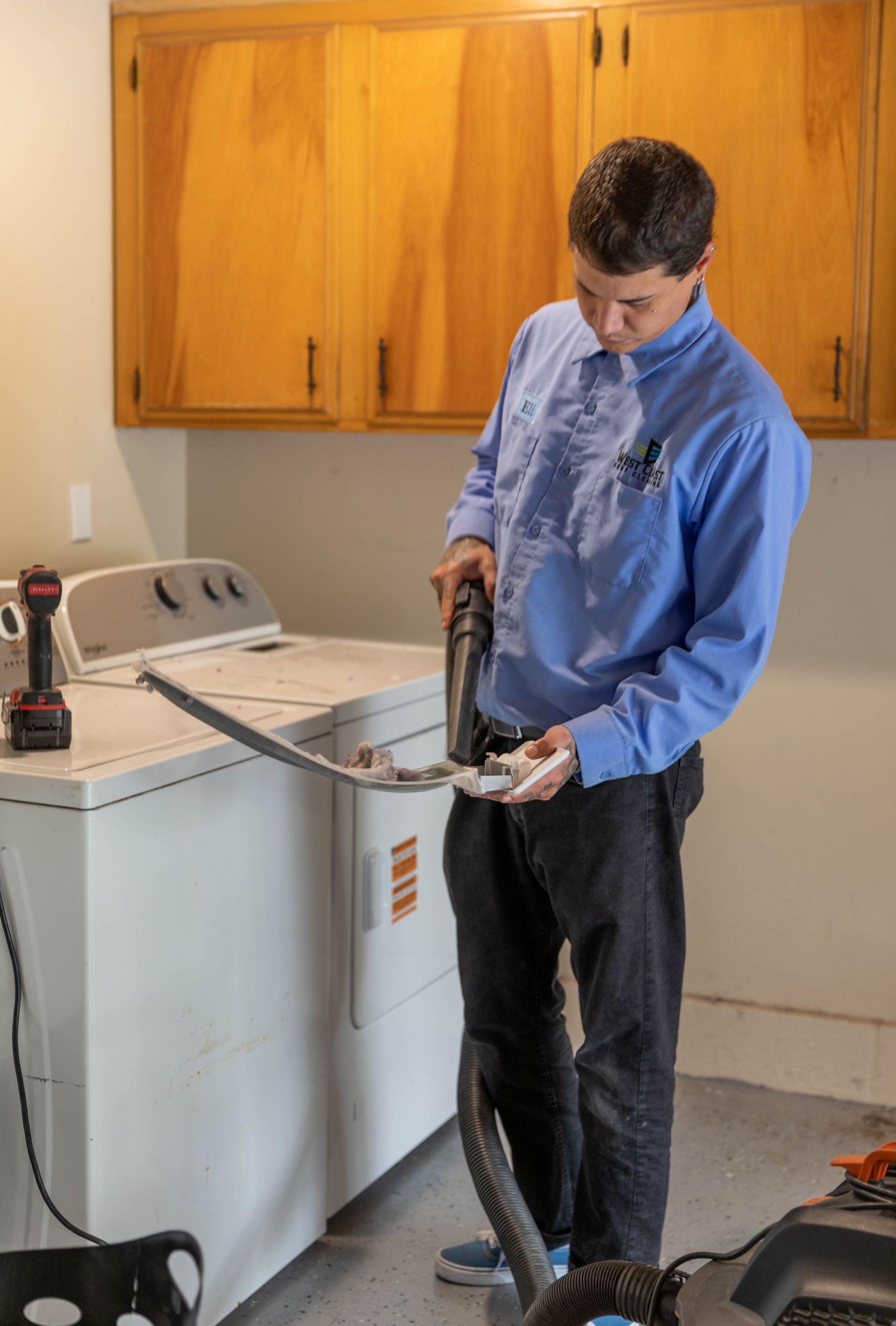 A person in a blue button-down shirt cleaning a dryer vent hose with a vacuum attachment in a laundry room.