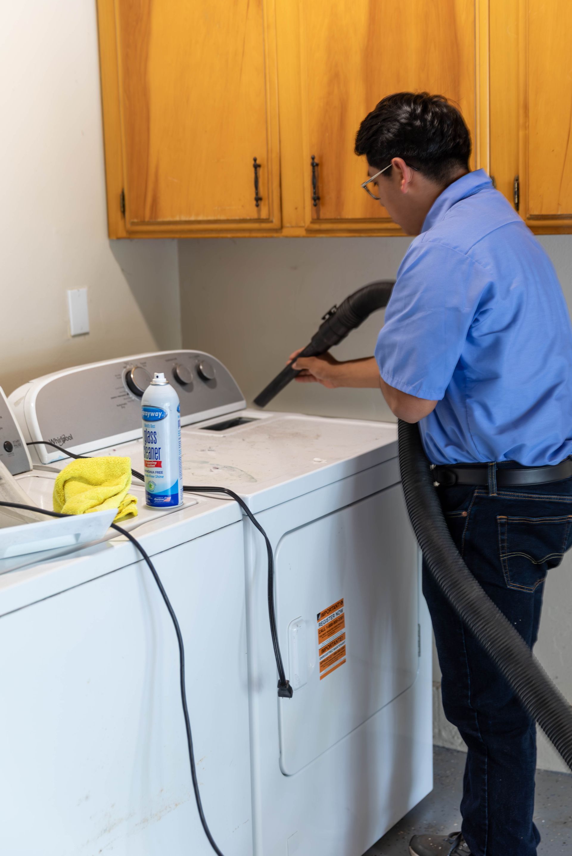 A person in a blue shirt cleans the top of a white clothes dryer with a vacuum hose in a laundry room with wood cabinets.