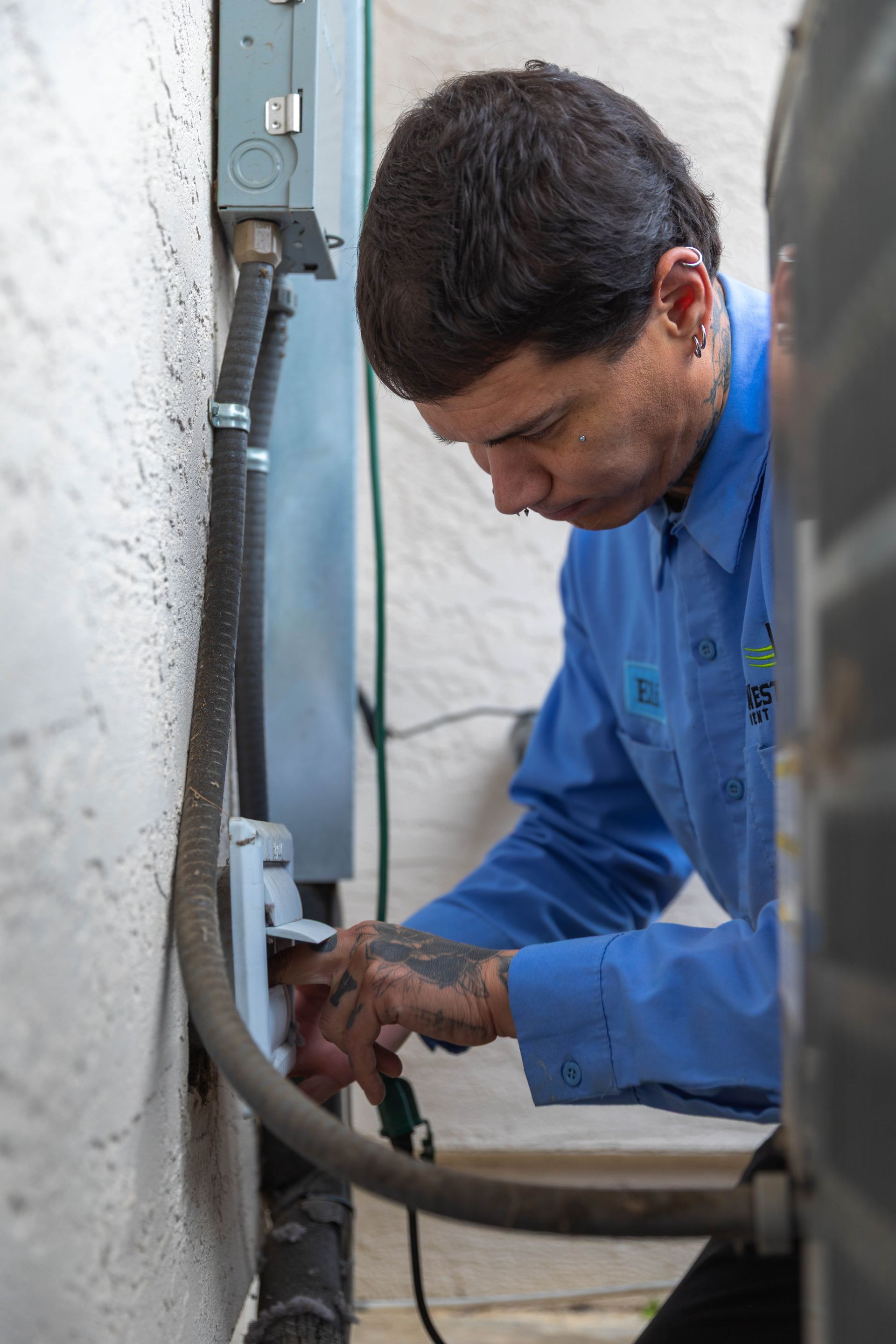 A technician in a blue uniform works on electrical wiring connected to an outdoor unit on a textured white wall.