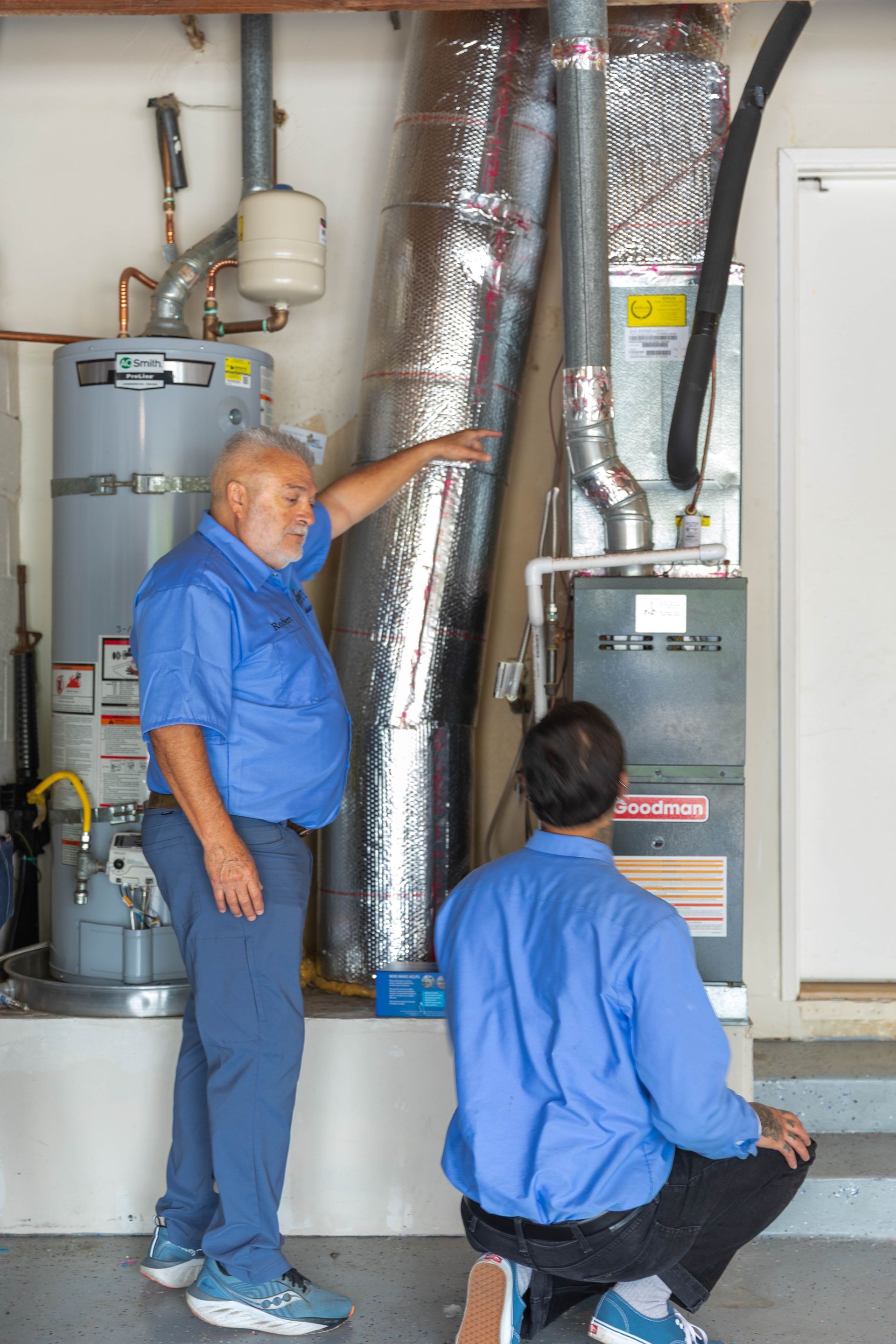 Two professionals in blue work shirts inspect a furnace and water heater setup in a utility room.