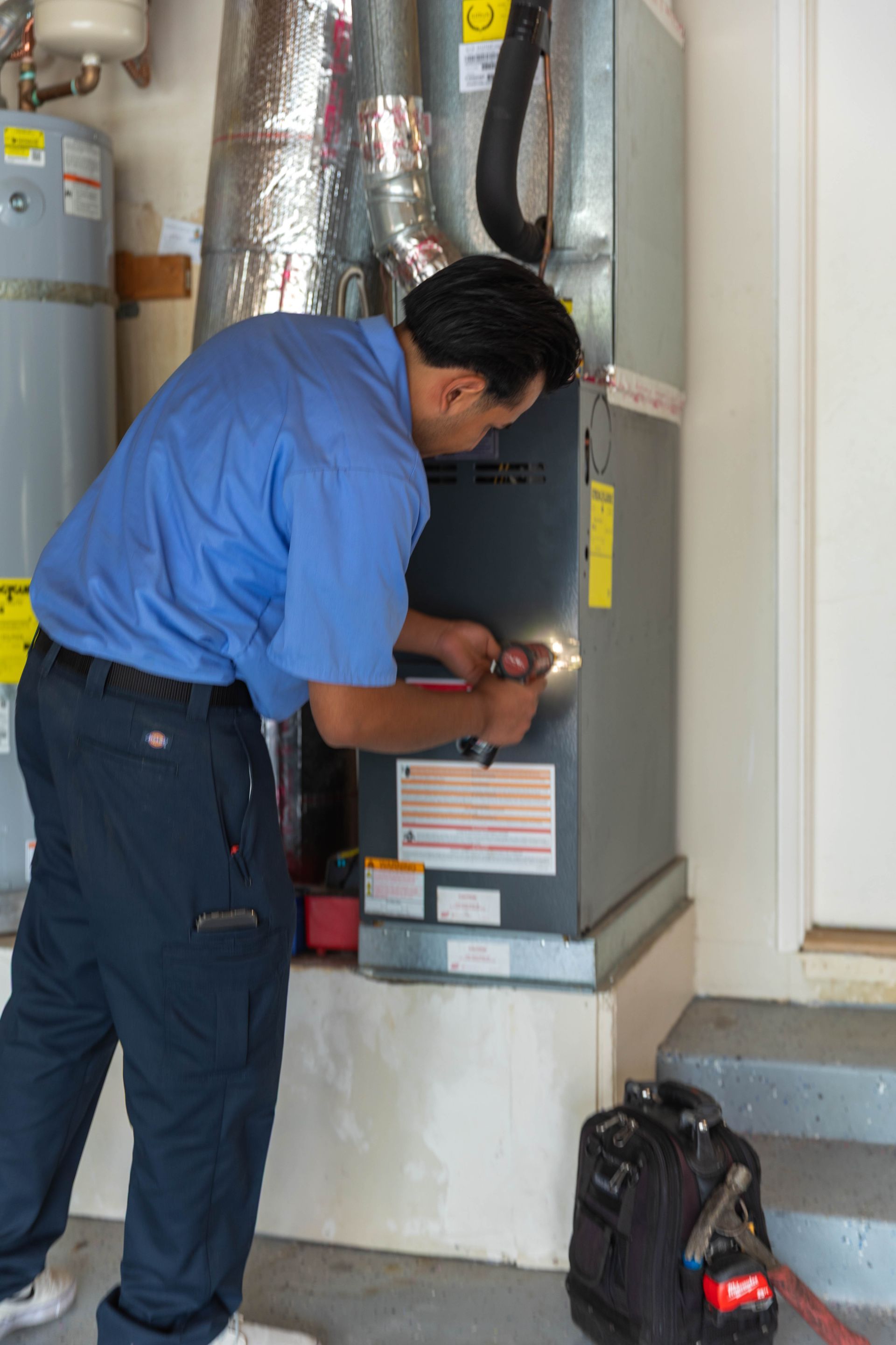 A technician in a blue uniform repairing a furnace in a residential utility room.