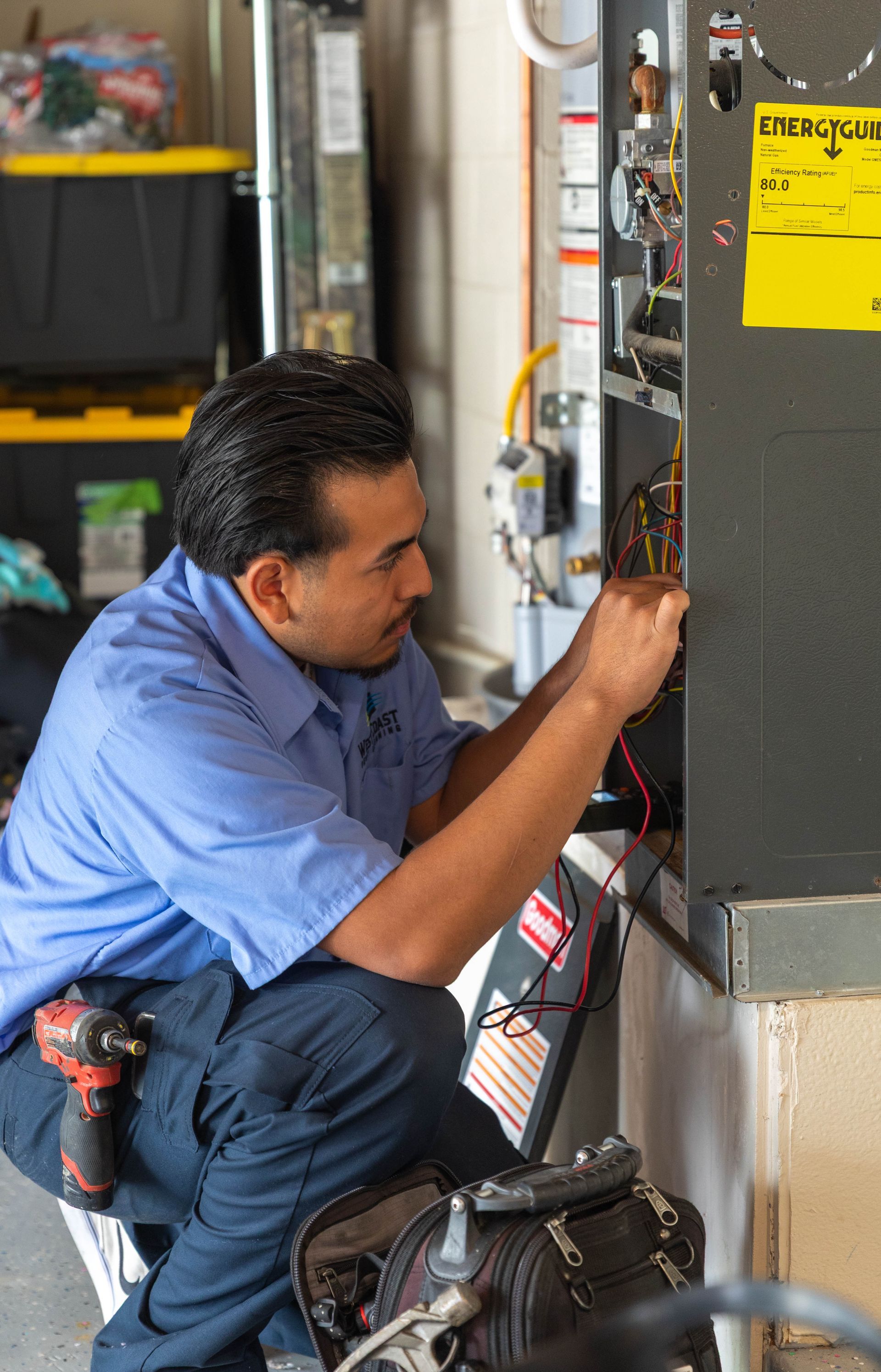 A technician in a blue uniform kneeling to repair a furnace, using tools to check the wiring inside the open unit.