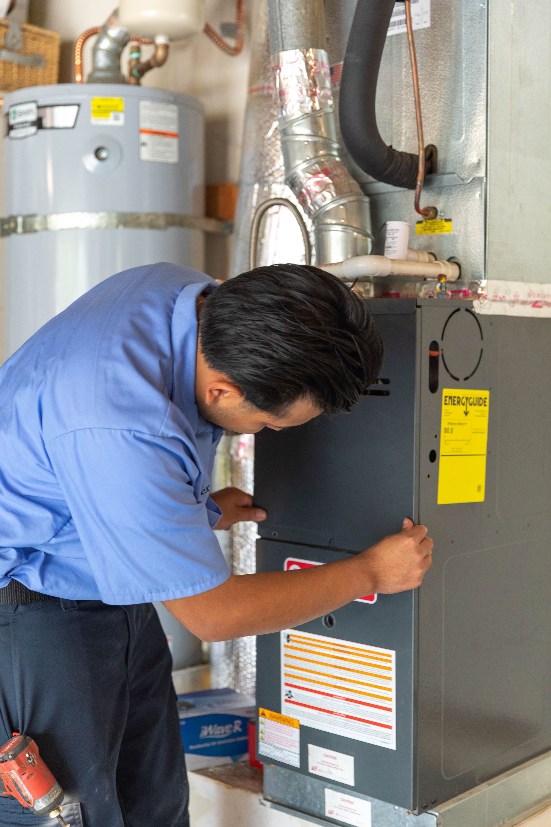 A technician in a blue uniform inspecting and servicing a furnace in a residential utility room.