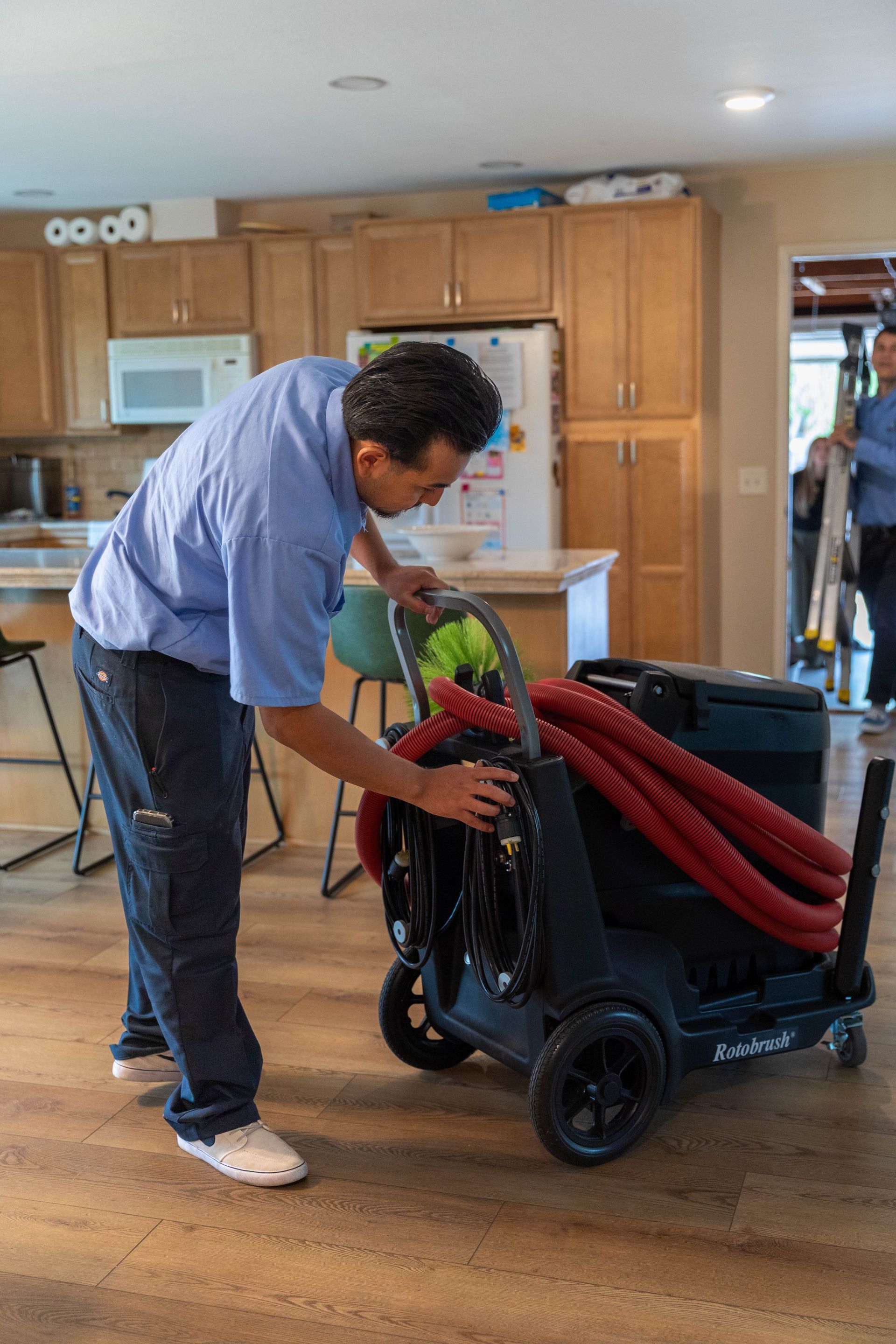 A person in a blue shirt adjusting a black carpet-cleaning machine with red hoses inside a home kitchen.