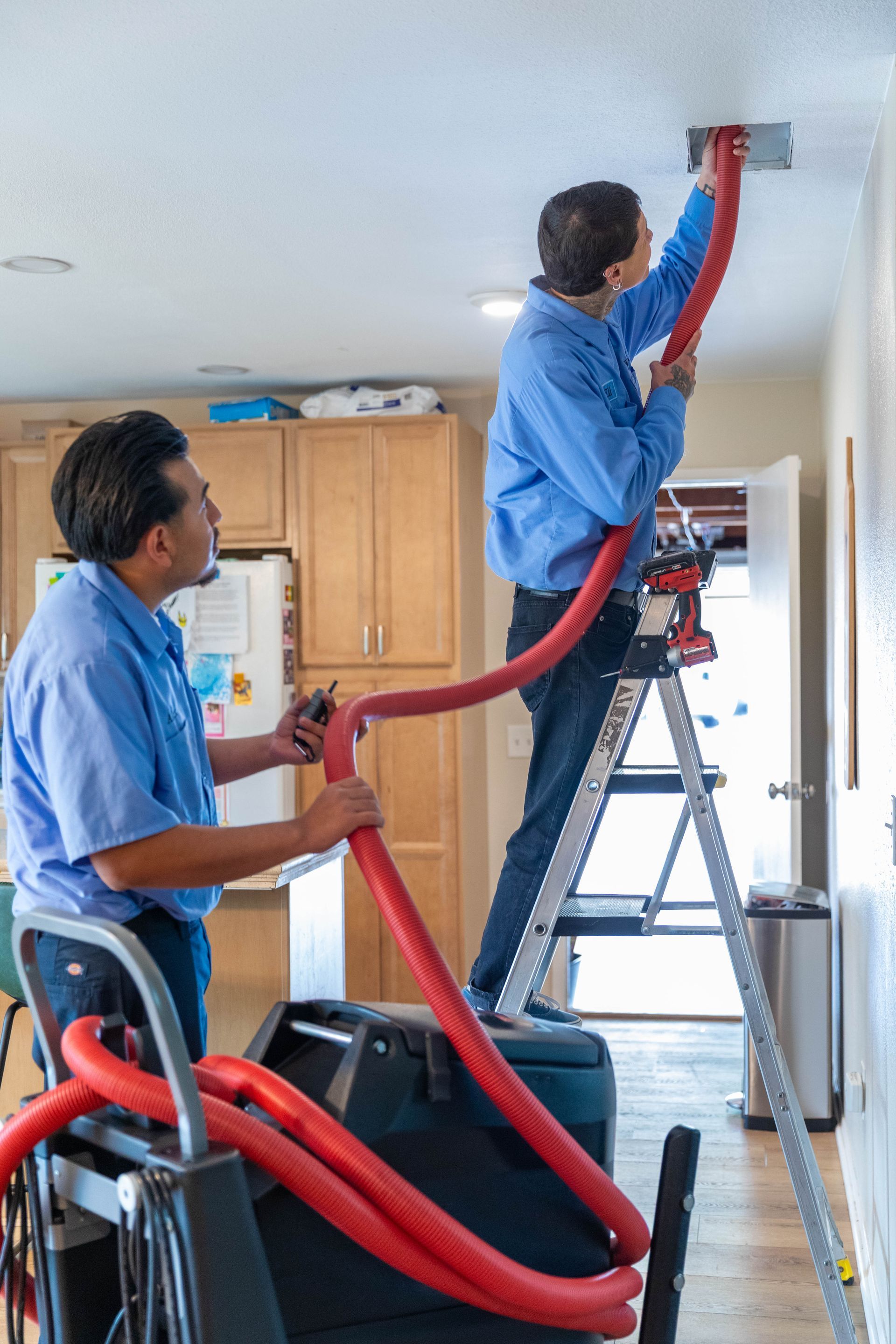 Two professionals in blue work shirts inspect a furnace and water heater setup in a utility room.