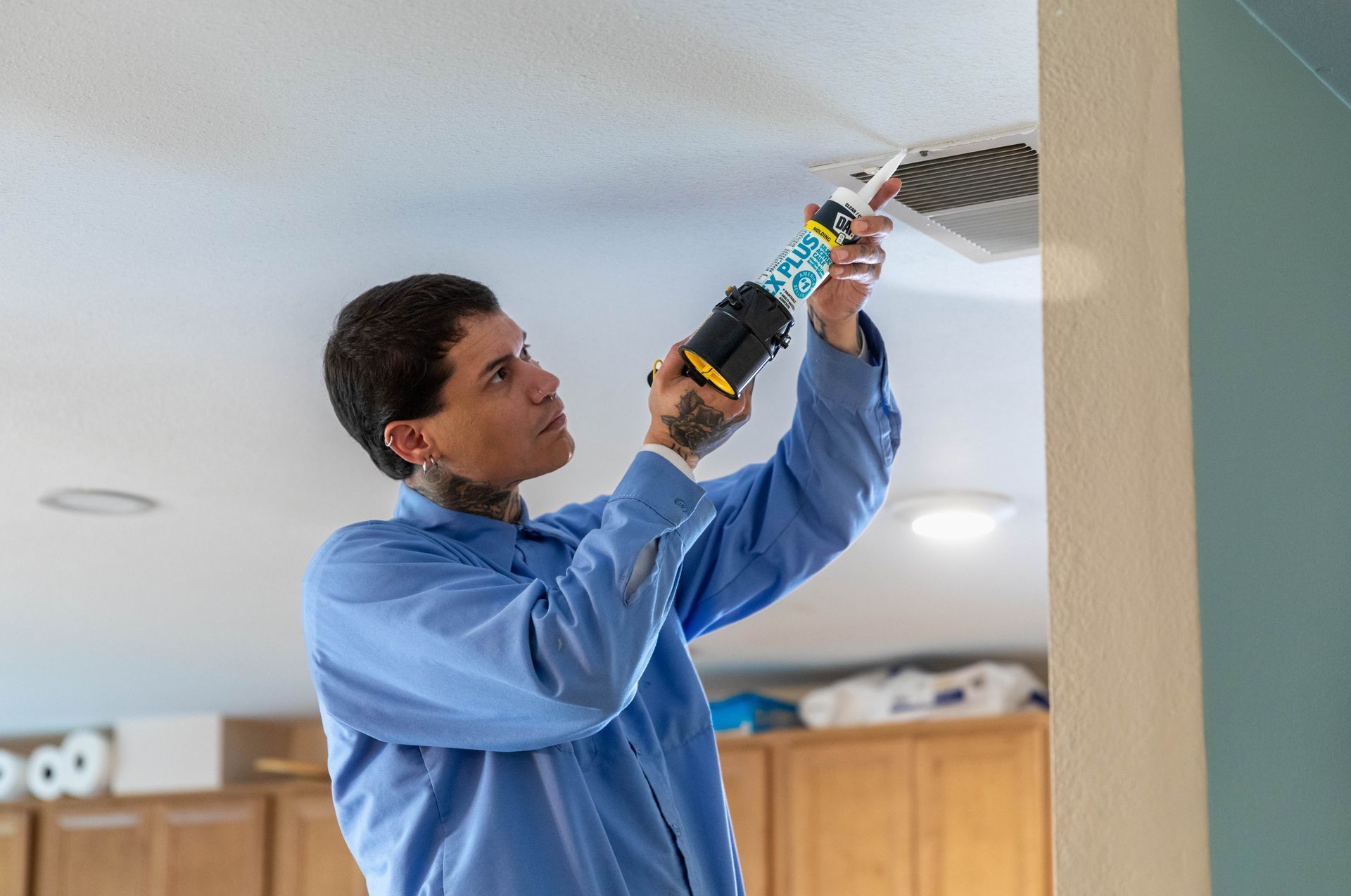 A technician in a blue shirt uses a caulk gun to apply sealant around a ceiling air vent.