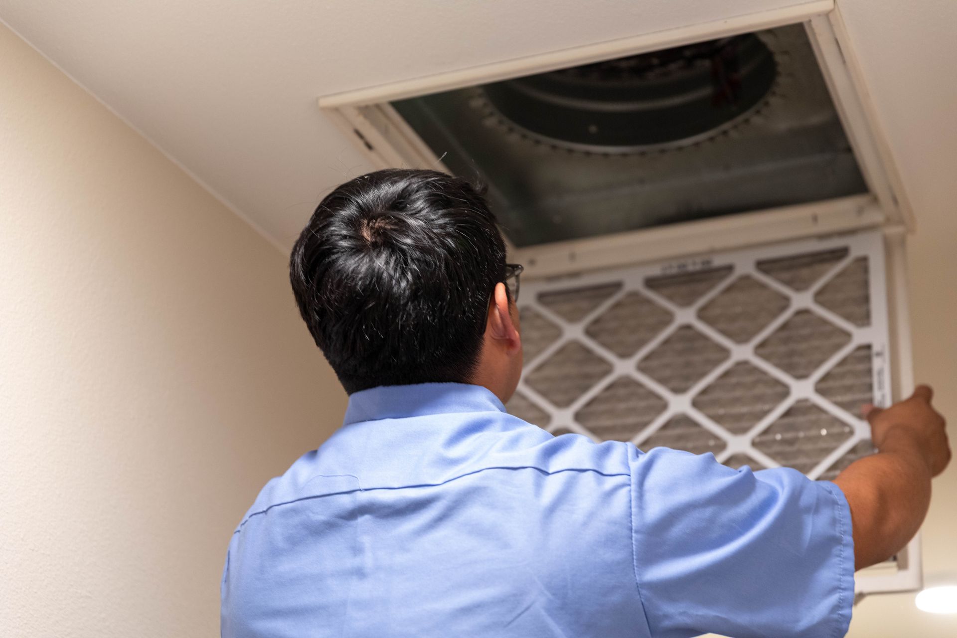 A technician in a light blue uniform replaces an air conditioning filter in a ceiling vent.