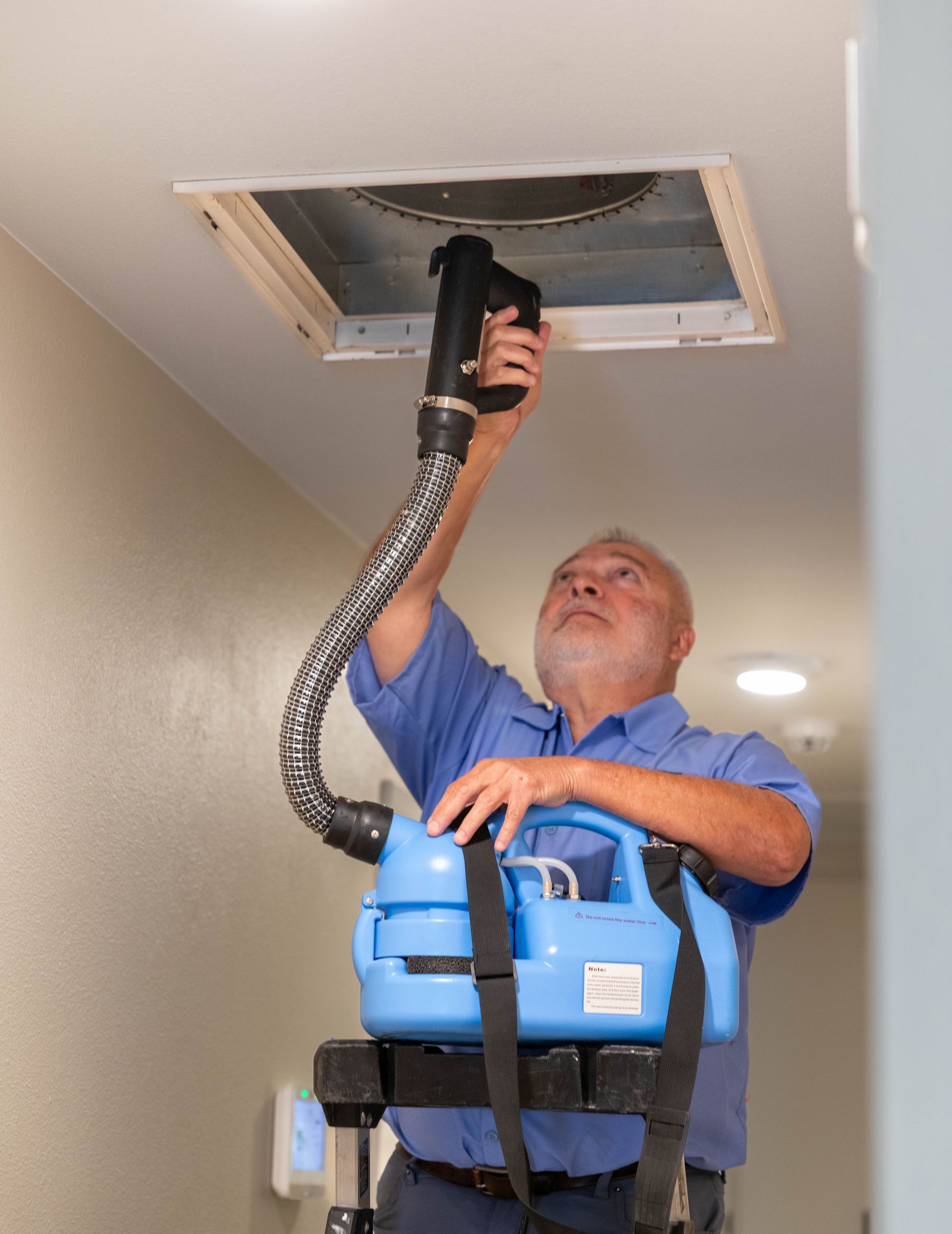 A technician uses a handheld device attached to a blue portable unit to clean a ceiling vent in a hallway.