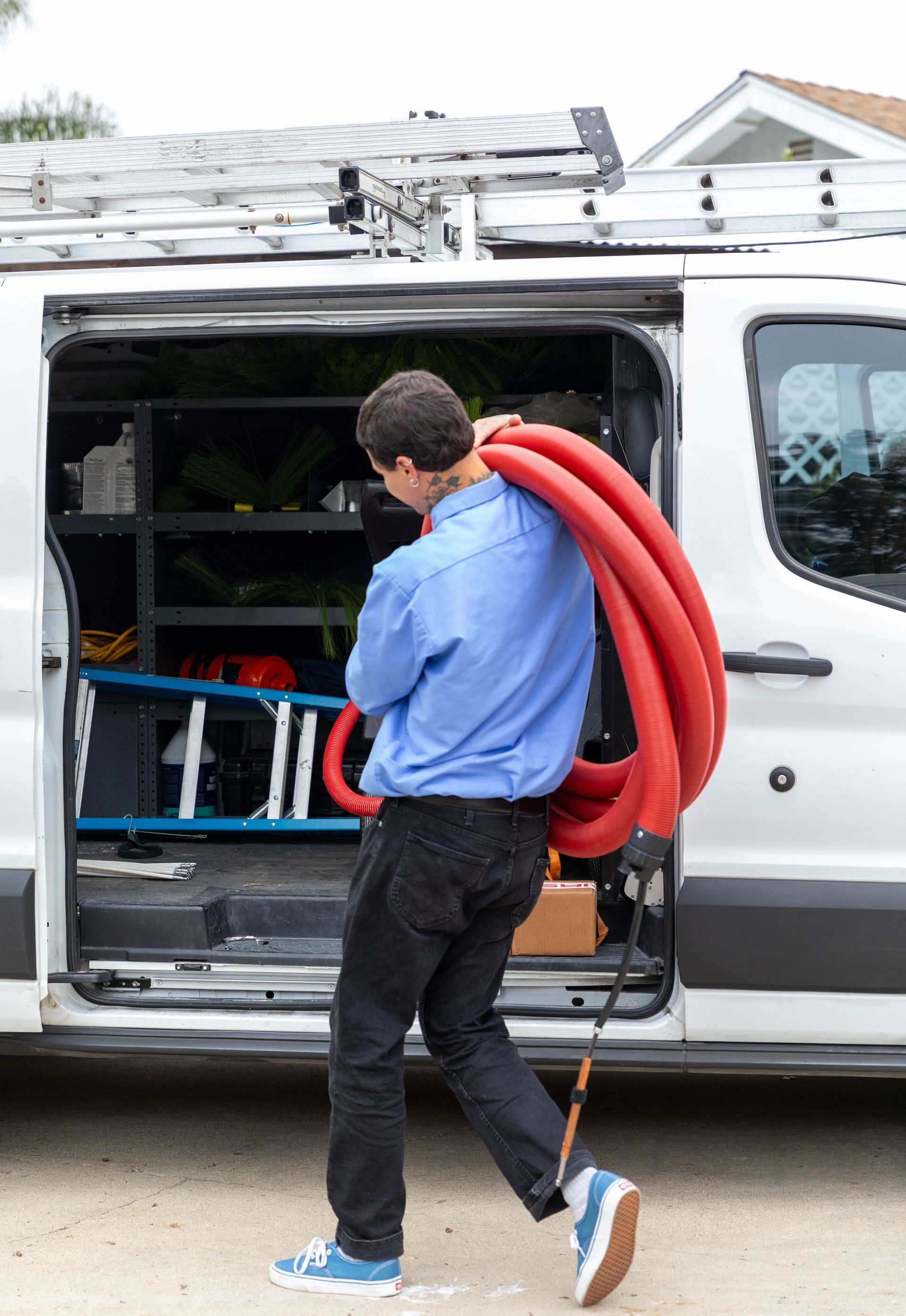 A technician in a blue shirt carries a large, red coiled hose away from a white service van with a roof ladder rack.