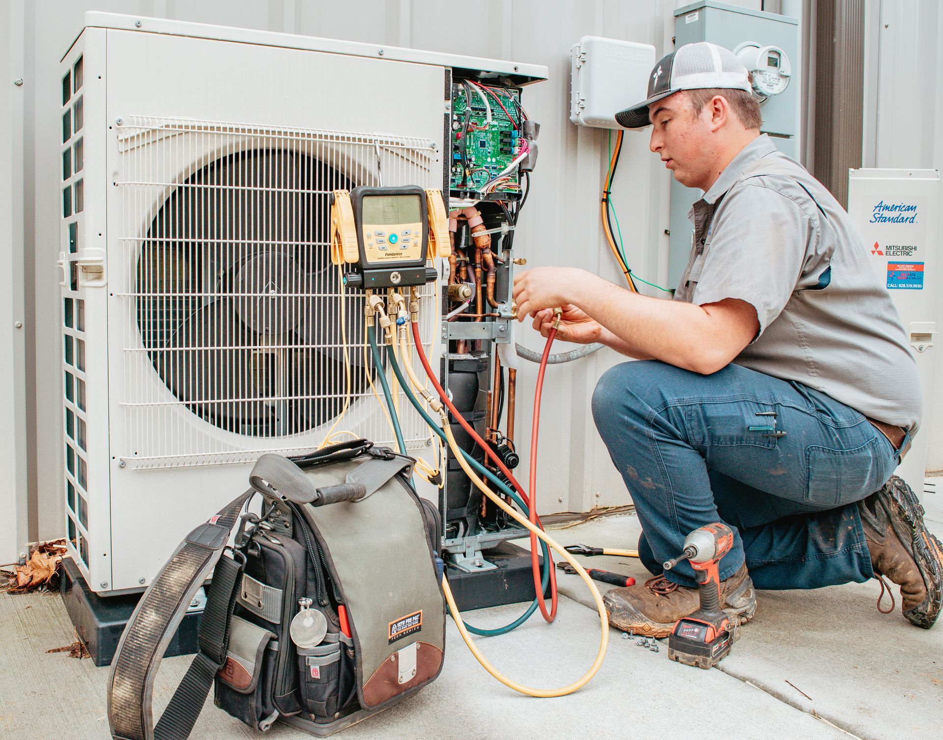 HVAC technician repairs an outdoor unit. He kneels, using gauges, tools, and colored hoses.