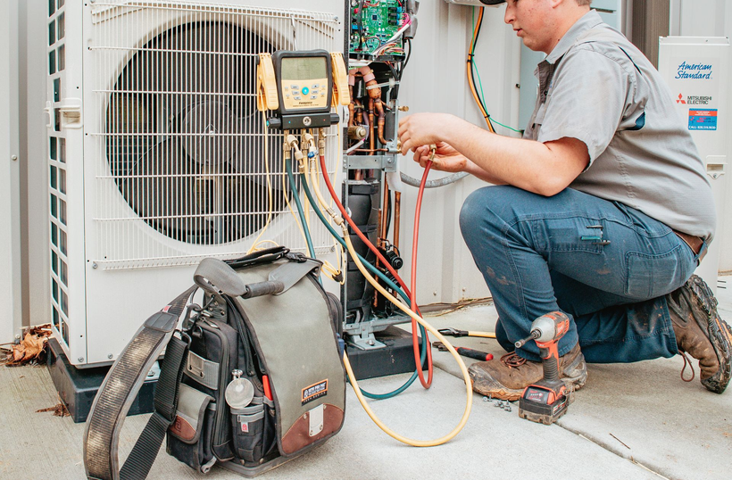 HVAC technician kneeling, working on outdoor air conditioning unit, using tools.