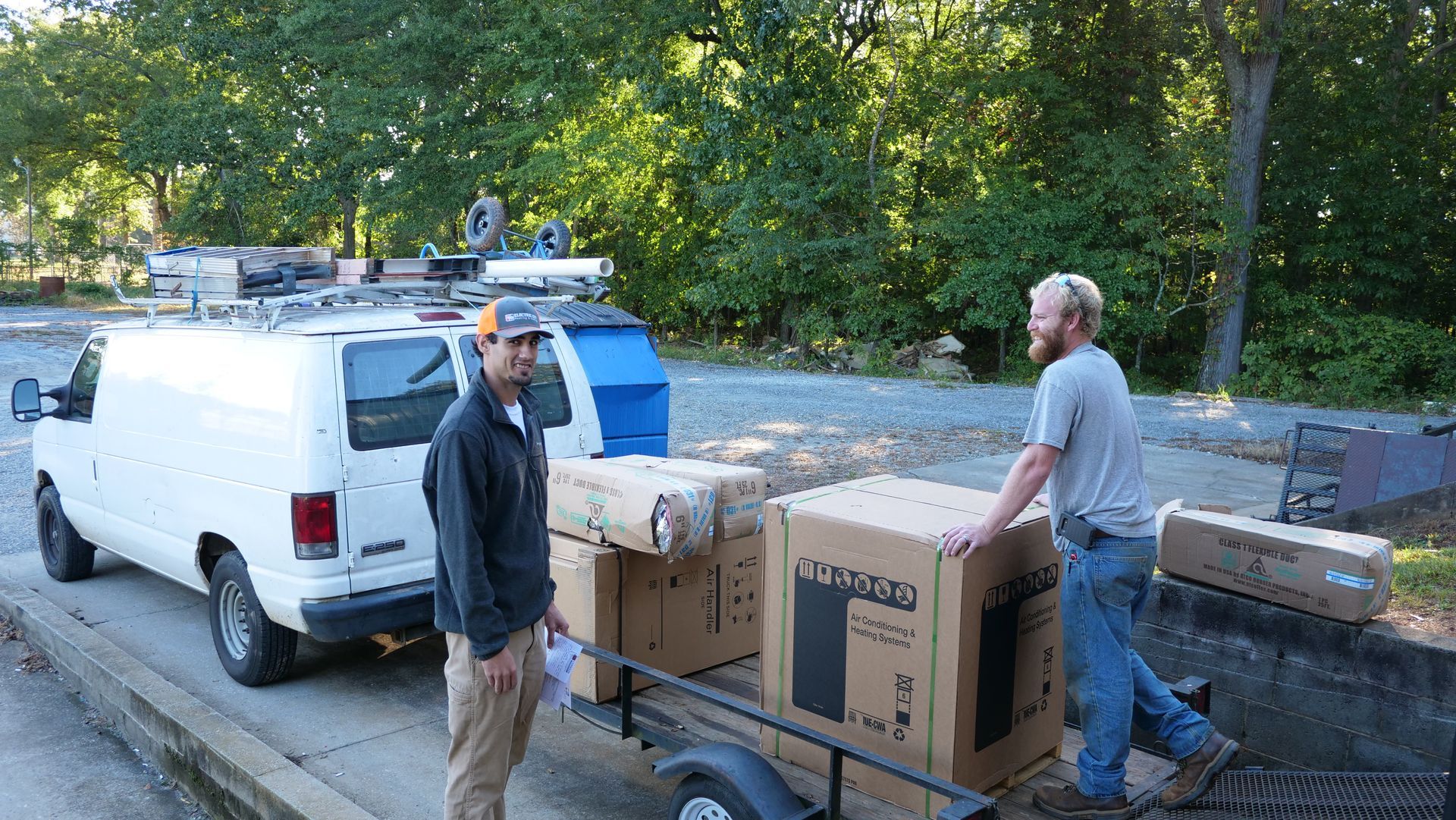 Two men loading boxes from a trailer next to a white van in front of trees.
