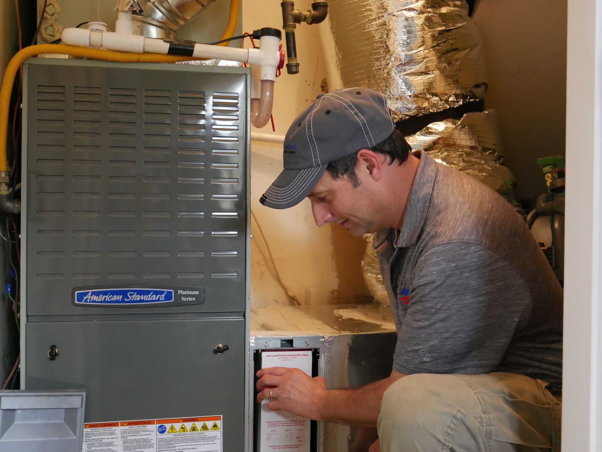 HVAC technician inspecting a furnace, kneeling beside it. Gray furnace, silver ductwork, wearing a cap.