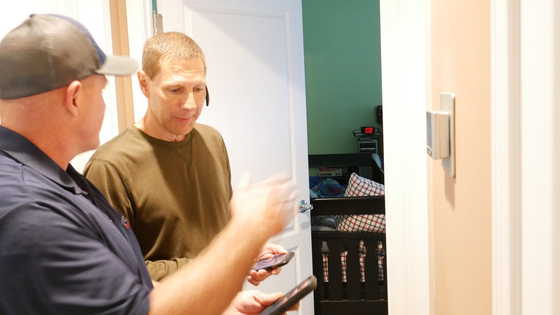 Two men looking at a smart thermostat on a wall. One gestures while holding a phone.