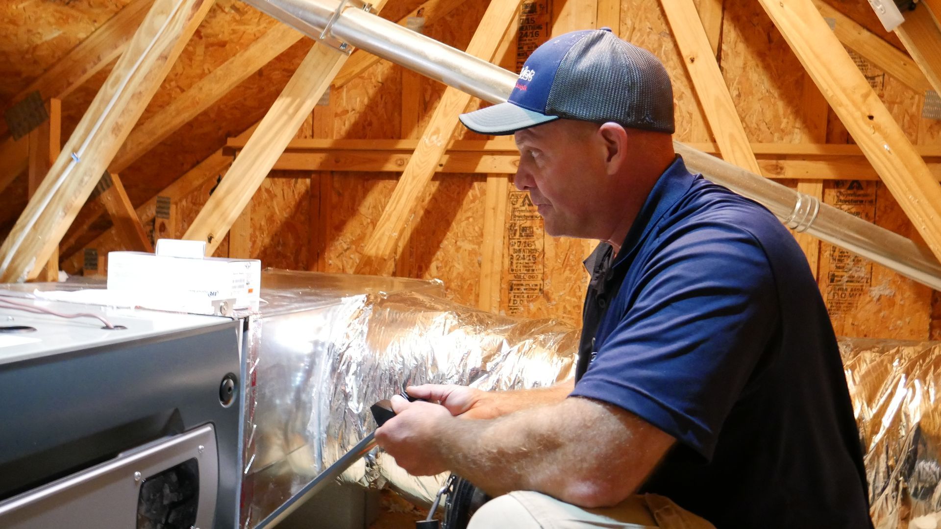 HVAC technician working on ductwork in an attic, wearing a hat and navy shirt.
