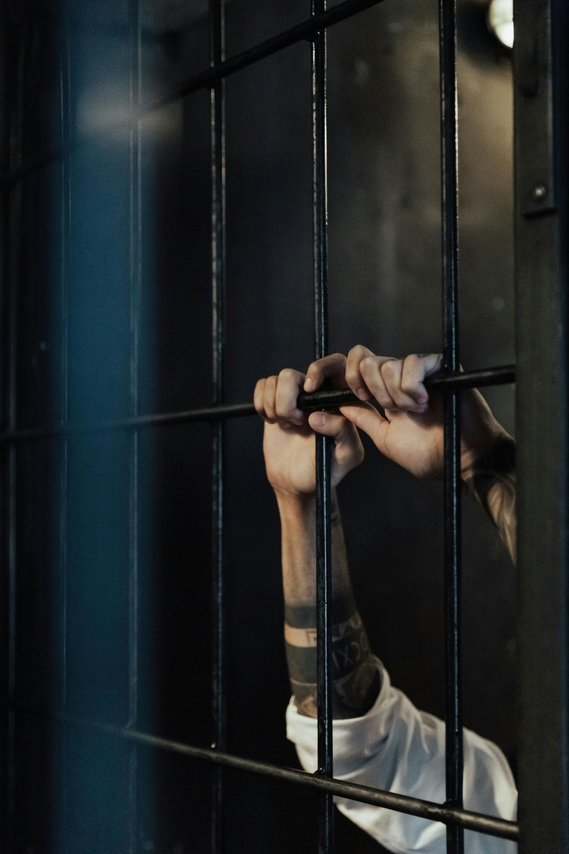 Person's arms holding prison bars. Tattooed forearms, white shirt. Dark room, bleak setting.