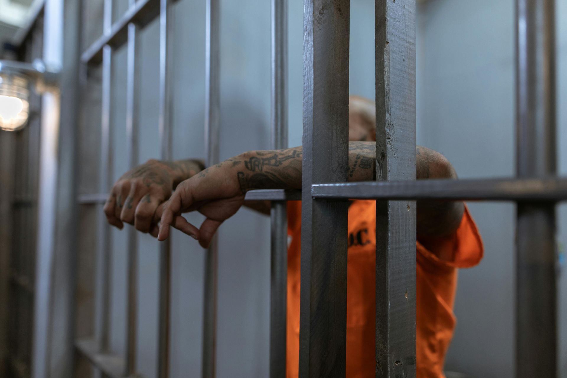 Person in orange prison uniform behind bars, hands on the bars, looking down.