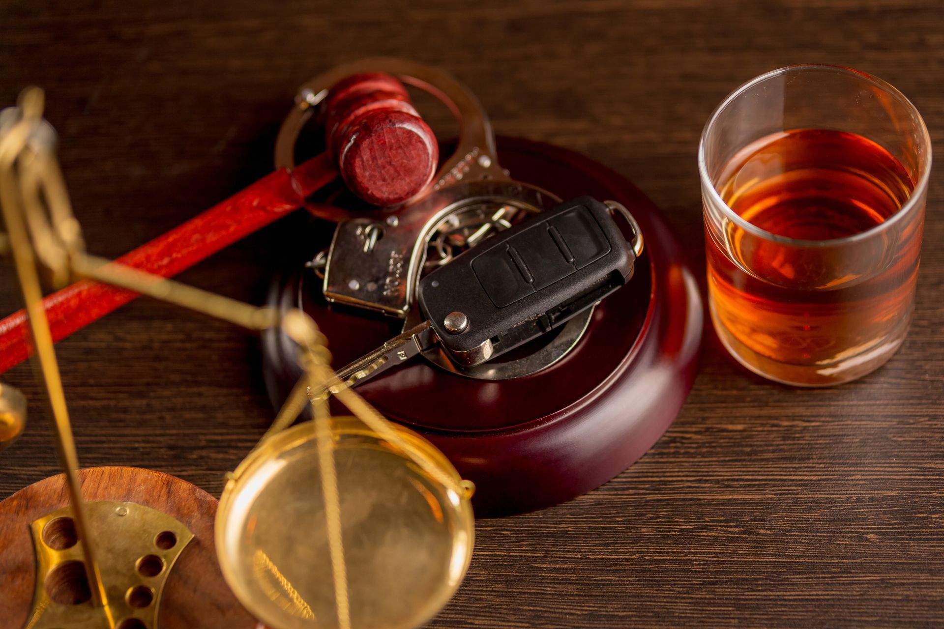 Scales of justice, gavel, handcuffs, car keys, and a drink on a wooden surface.
