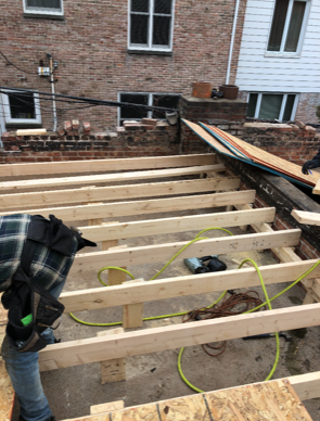 Un homme travaille sur une terrasse en bois devant un bâtiment en briques.