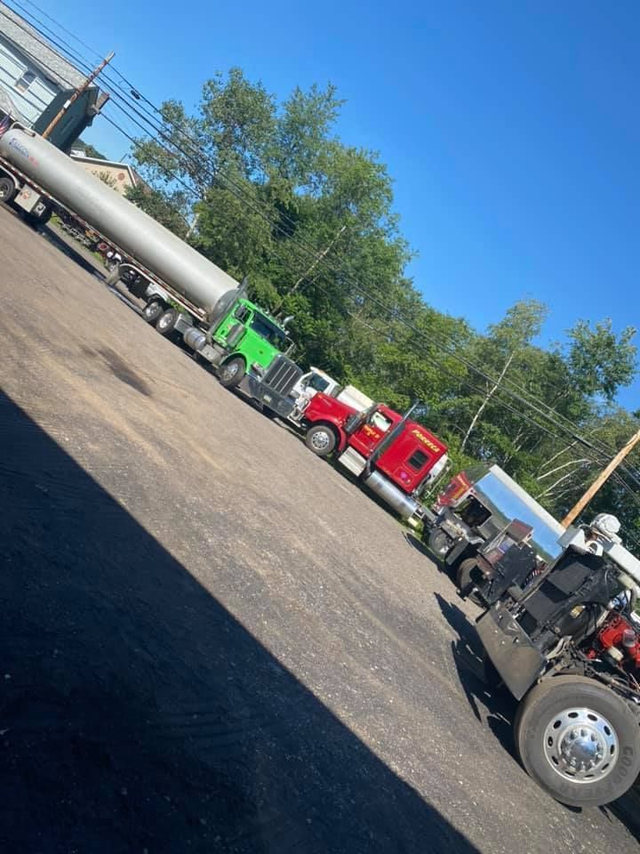 Three semi-trucks of different colors parked on an asphalt lot on a sunny day. One hauls a long, silver cylinder.