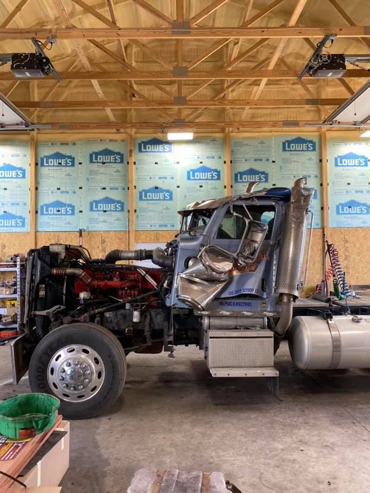Blue semi-truck cab in a garage with its engine exposed. Wood beams and Lowe's insulation are in the background.