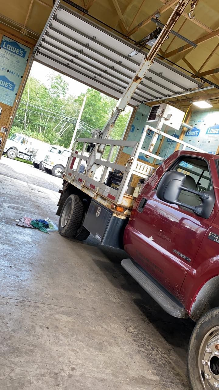 Red truck parked inside a building, white machinery in the truck bed, open bay door reveals trees.