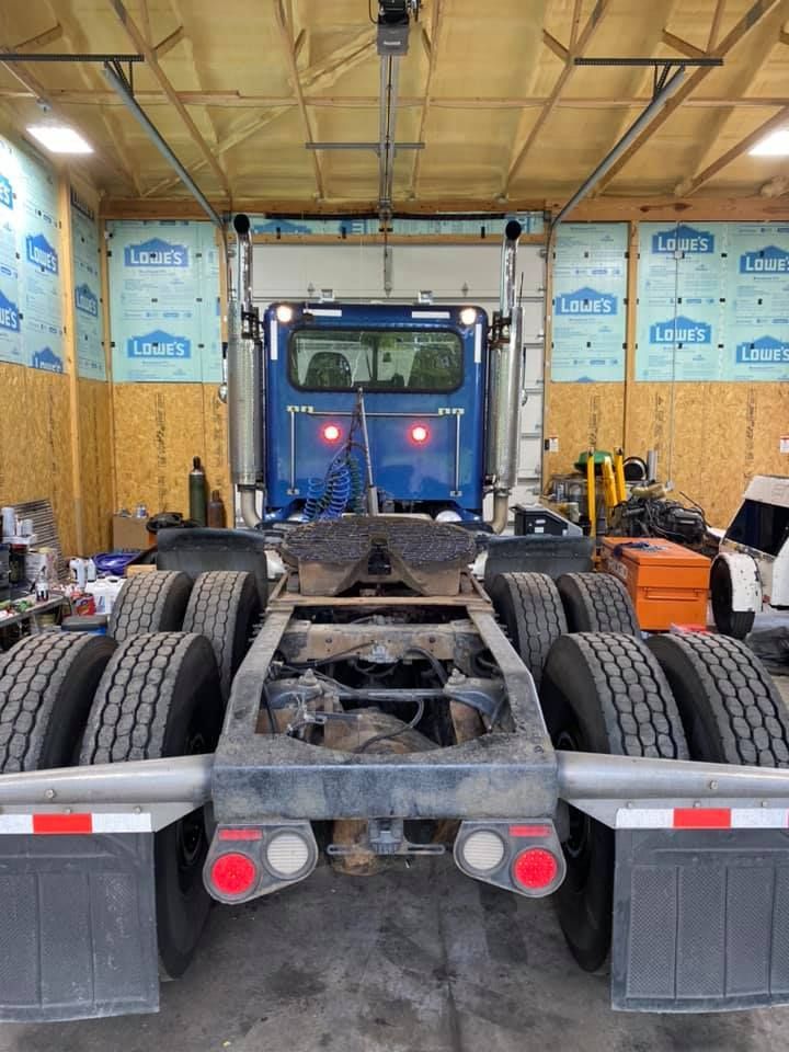 Back of a blue semi-truck cab and chassis inside a garage. Rear tires, taillights, and exhaust pipes are visible.