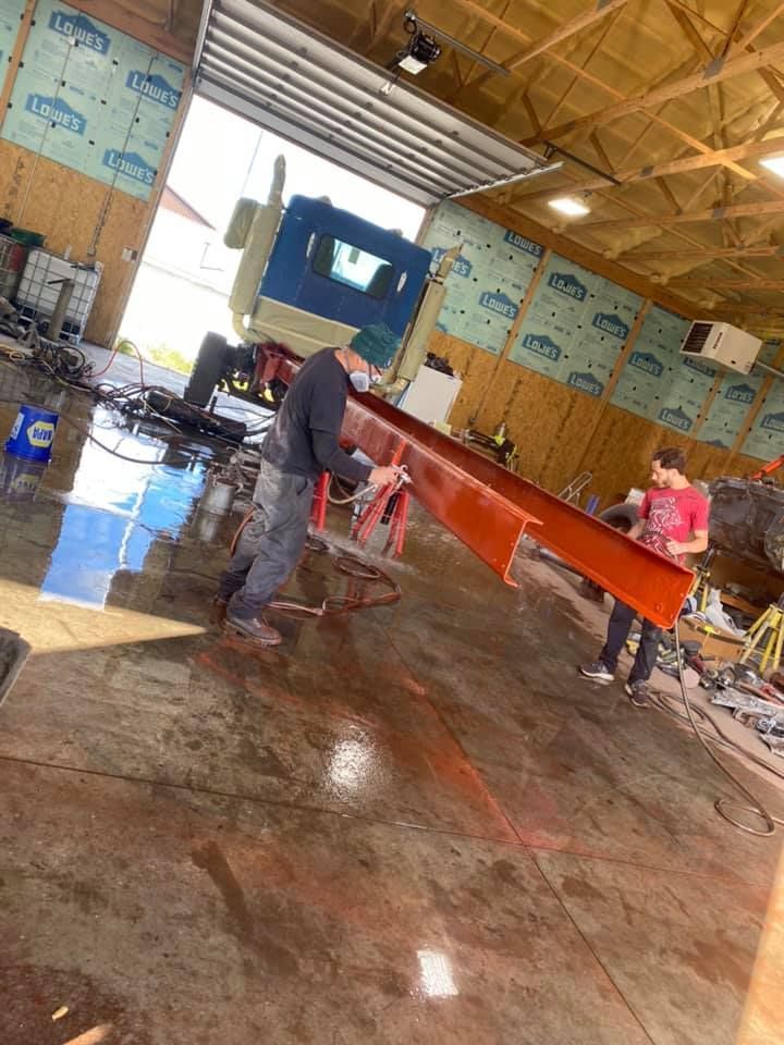 Two men spray painting an orange metal beam inside a workshop with a truck visible in the background.